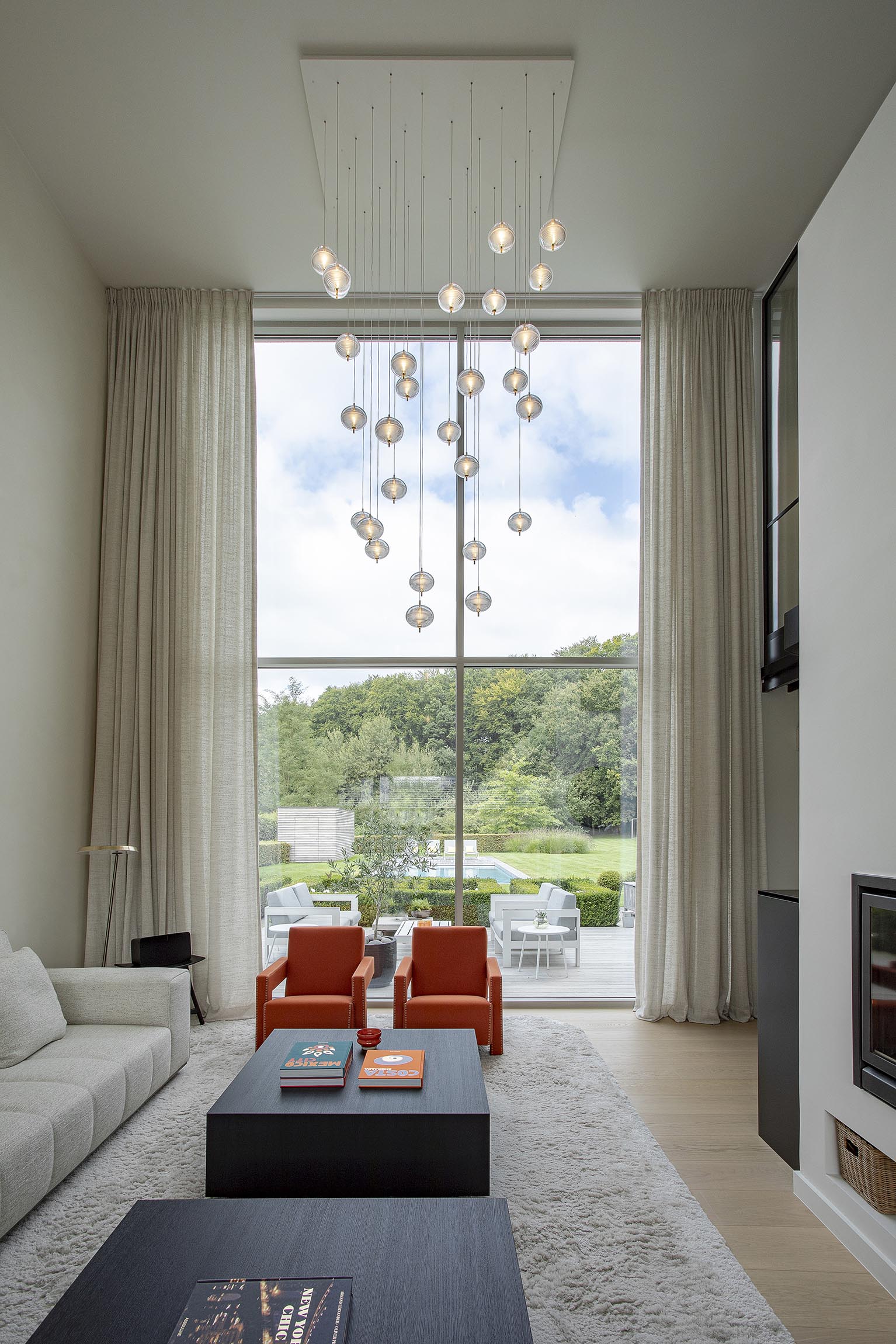 Salon contemporain haut de gamme avec grande baie vitrée, fauteuils orange et suspension sculpturale en verre soufflé