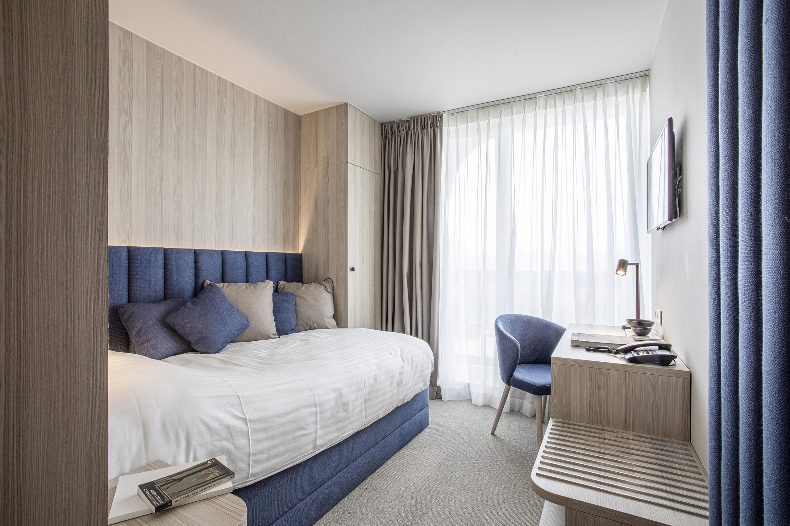 Chambre d’hôtel contemporaine aux 3 Clés à Gembloux, avec tête de lit bleue sur mesure, habillage mural en bois clair et éclairage indirect chaleureux