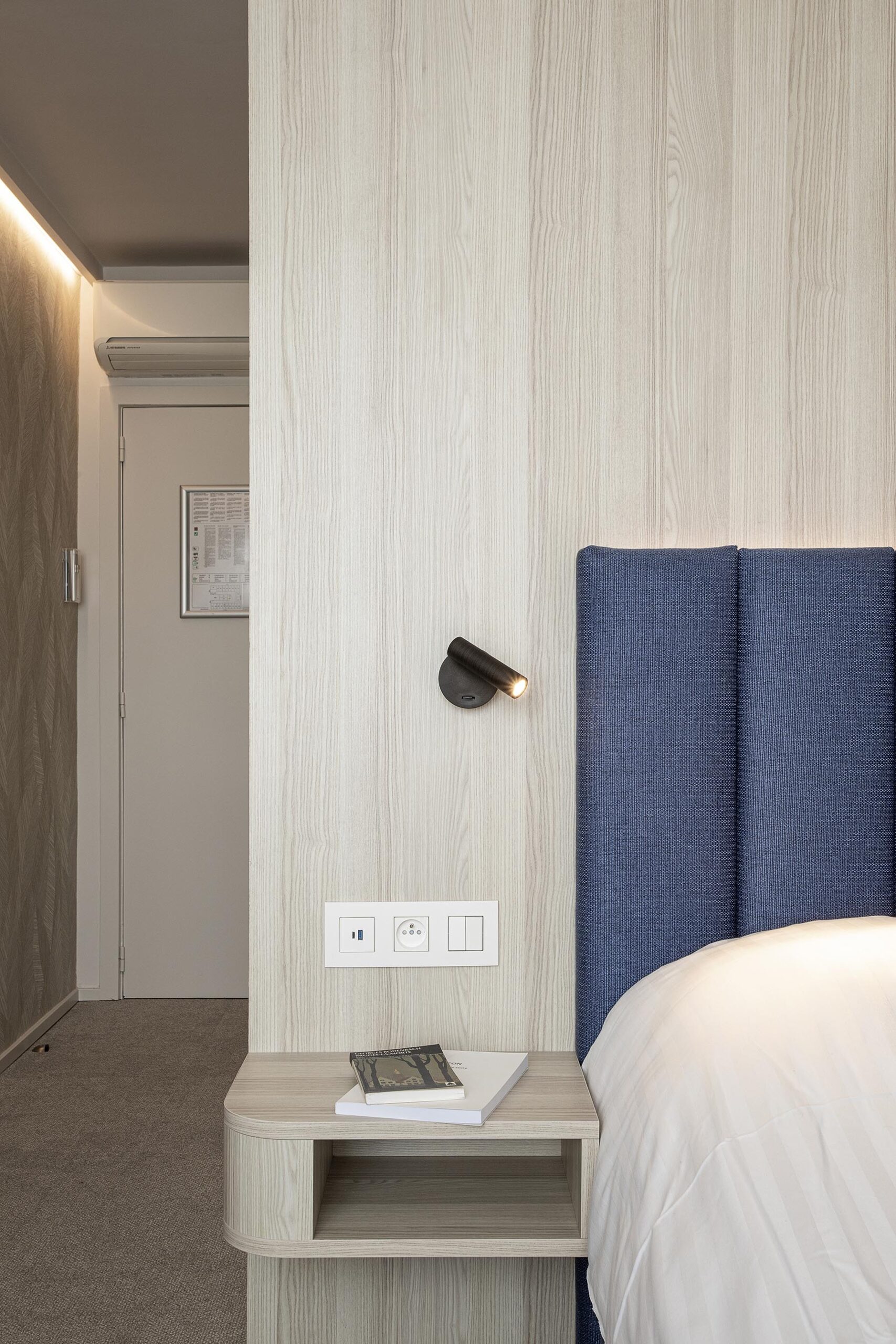 Chambre d’hôtel contemporaine aux 3 Clés à Gembloux, avec tête de lit bleue sur mesure, habillage mural en bois clair et éclairage indirect chaleureux.