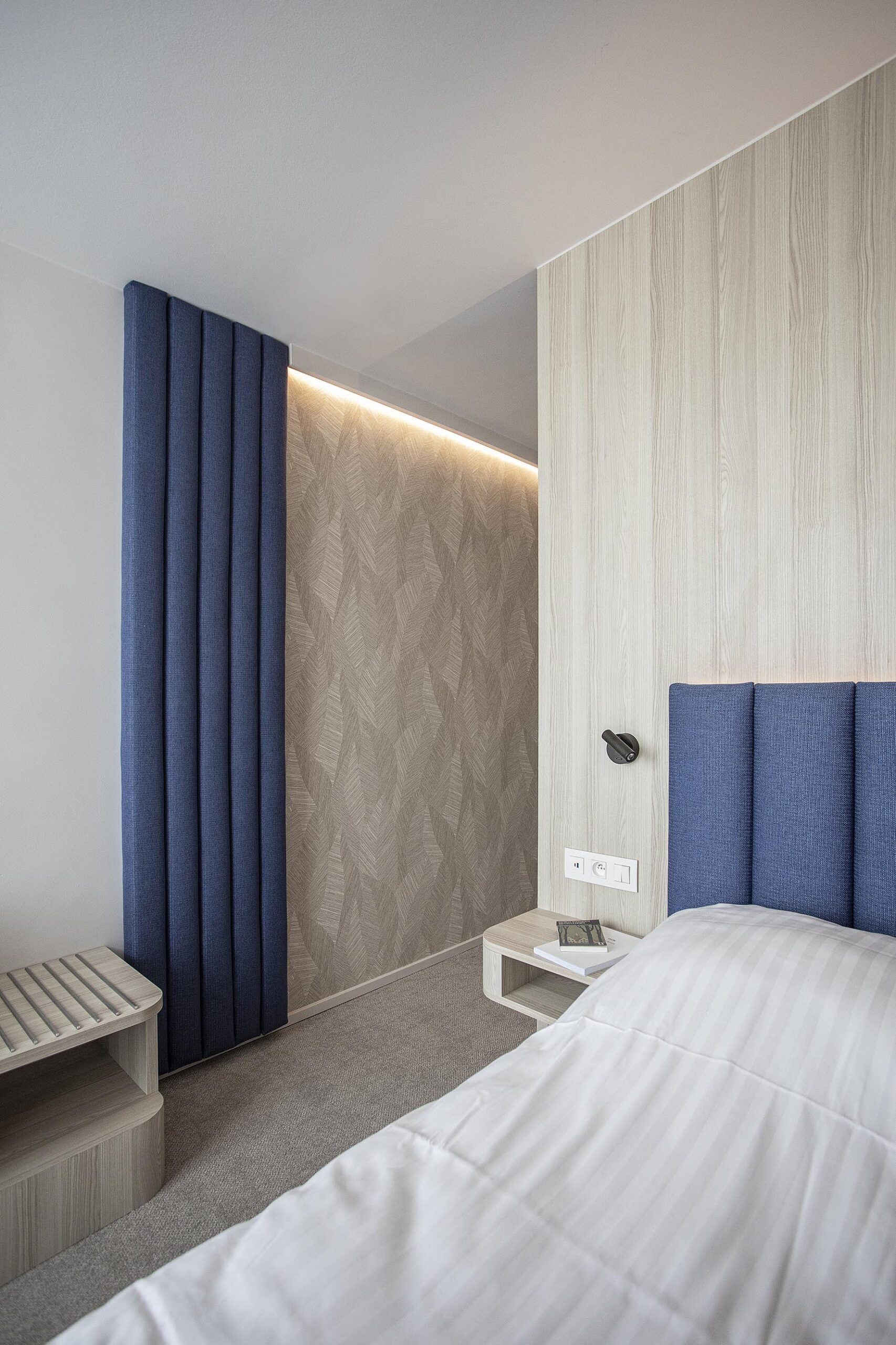 Chambre d’hôtel contemporaine aux 3 Clés à Gembloux, avec tête de lit bleue sur mesure, habillage mural en bois clair et éclairage indirect chaleureux.