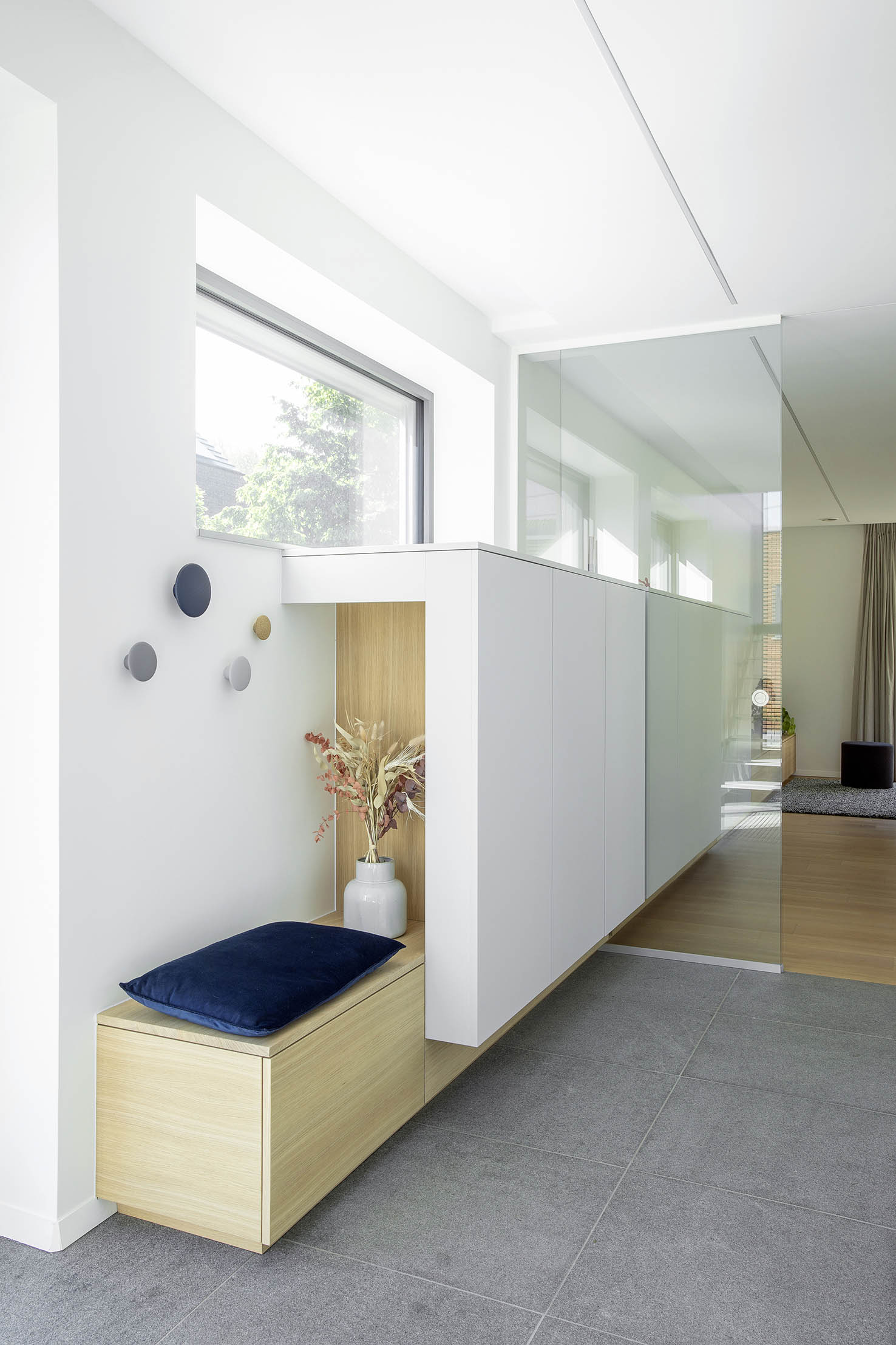 Hall d’entrée contemporain avec mobilier sur mesure en bois clair, banquette intégrée et paroi vitrée, projet d’architecture d’intérieur à Arlon.