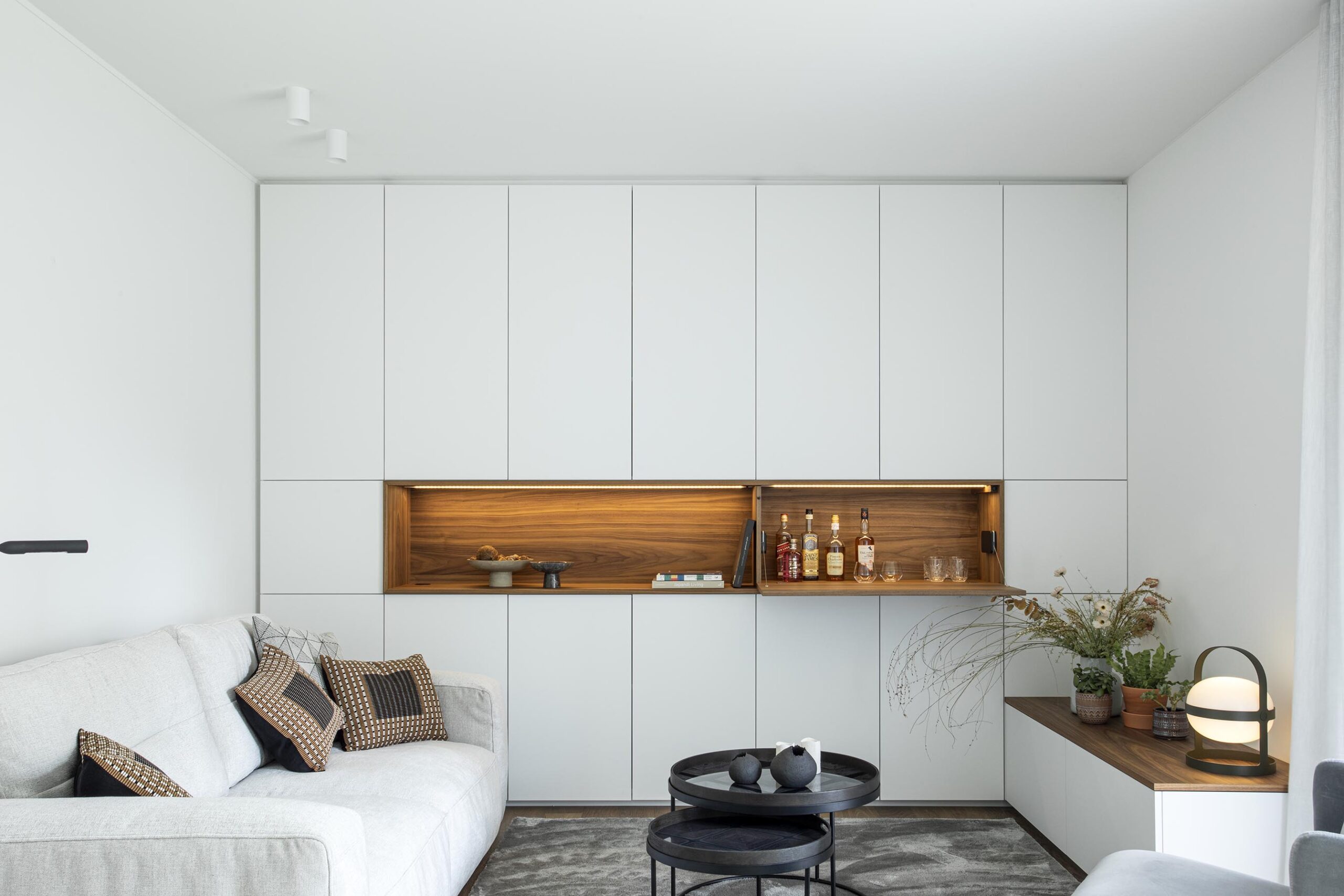 Salon contemporain lumineux avec mobilier sur mesure blanc et bois, niche intégrée éclairée et grandes baies vitrées, projet d’architecture d’intérieur à Arlon