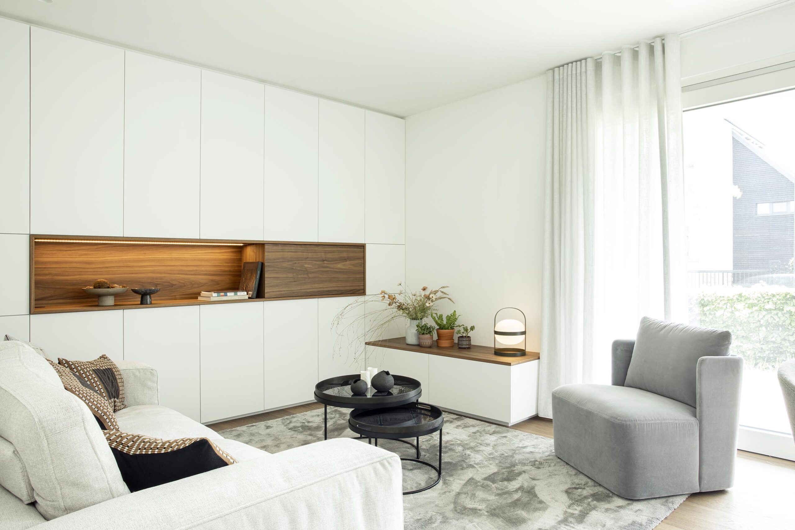 Salon contemporain lumineux avec mobilier sur mesure blanc et bois, niche intégrée éclairée et grandes baies vitrées, projet d’architecture d’intérieur à Arlon