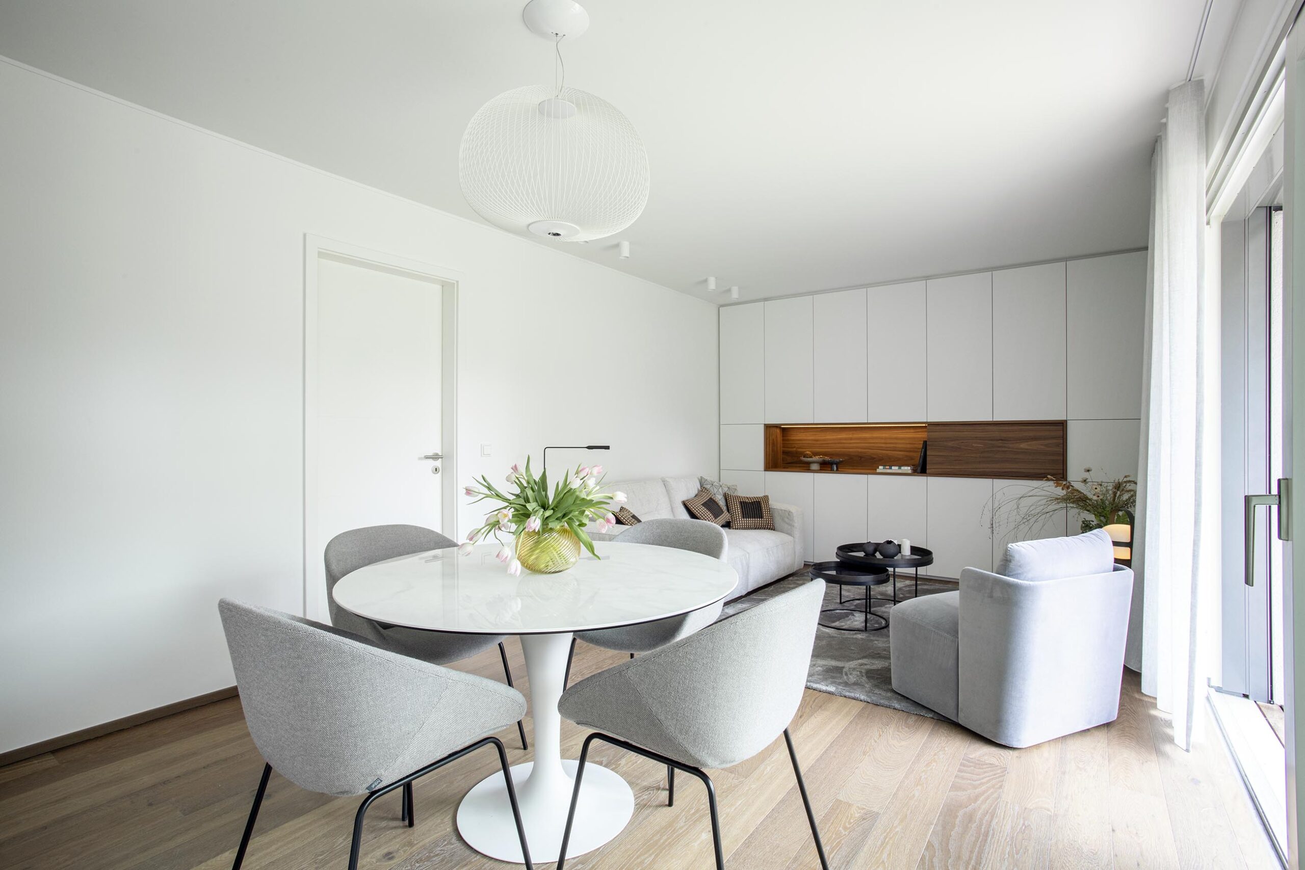 Salon contemporain lumineux avec mobilier sur mesure blanc et bois, niche intégrée éclairée et grandes baies vitrées, projet d’architecture d’intérieur à Arlon